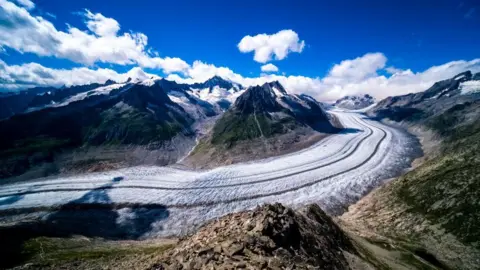 Getty Images View of the Aletsch Glacier. Ice sweeps round from right to left, with mountains either side. Above is a blue sky with some clouds.